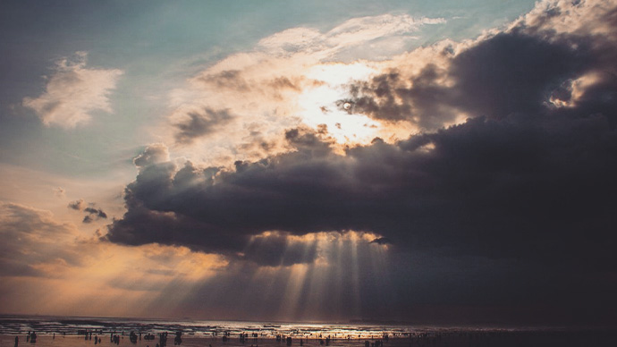 Sun peeking through clouds, sun rays shining down on ocean, people on the beach 