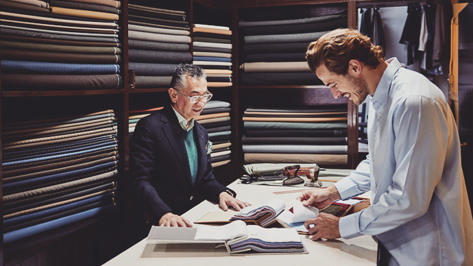 Medium wide shot of smiling client looking through fabric samples while ordering a custom suit in tailors workshop