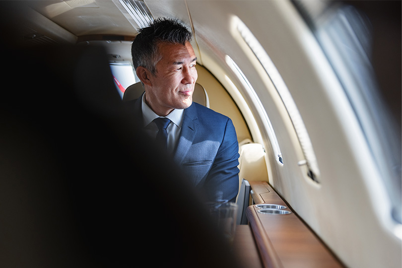 Mature Asian businessman in a suit looking out of the window in a luxurious private jet, reflecting on his success and aspirations.