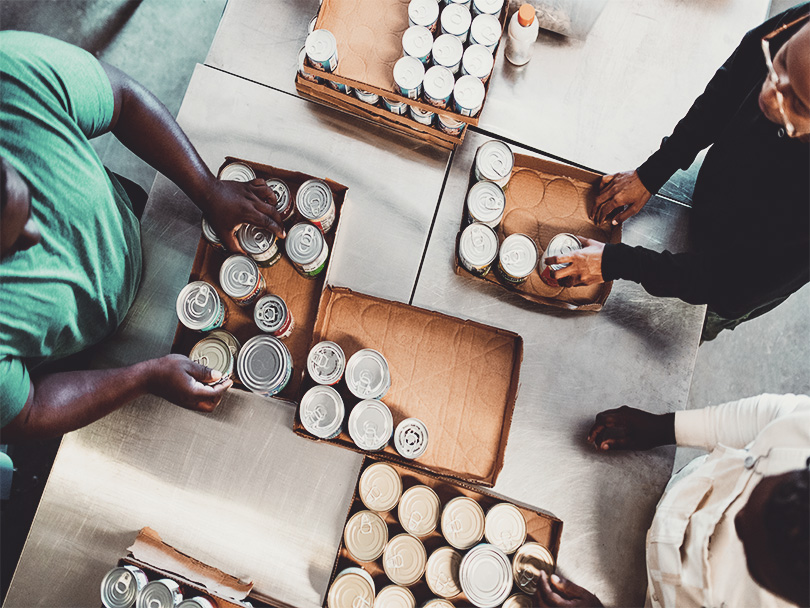 A group of young adult friends volunteer time working at a food bank, processing donations of packaged food products and clothing.  Overhead view of them sorting canned food.